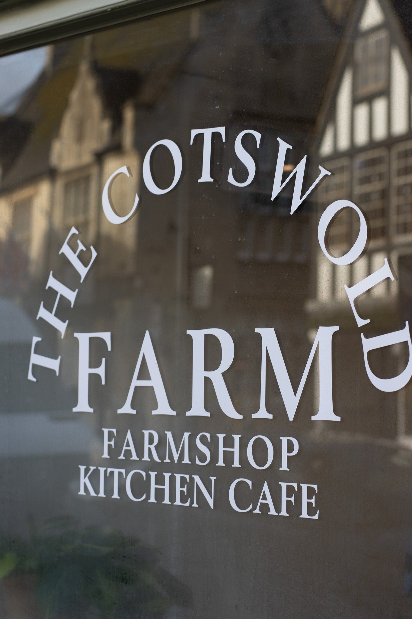 Close-up of a glass window with white text that reads "FARMSHOP, KITCHEN, CAFE." Reflection of buildings can be seen on the glass. The Cotswold Farm.
