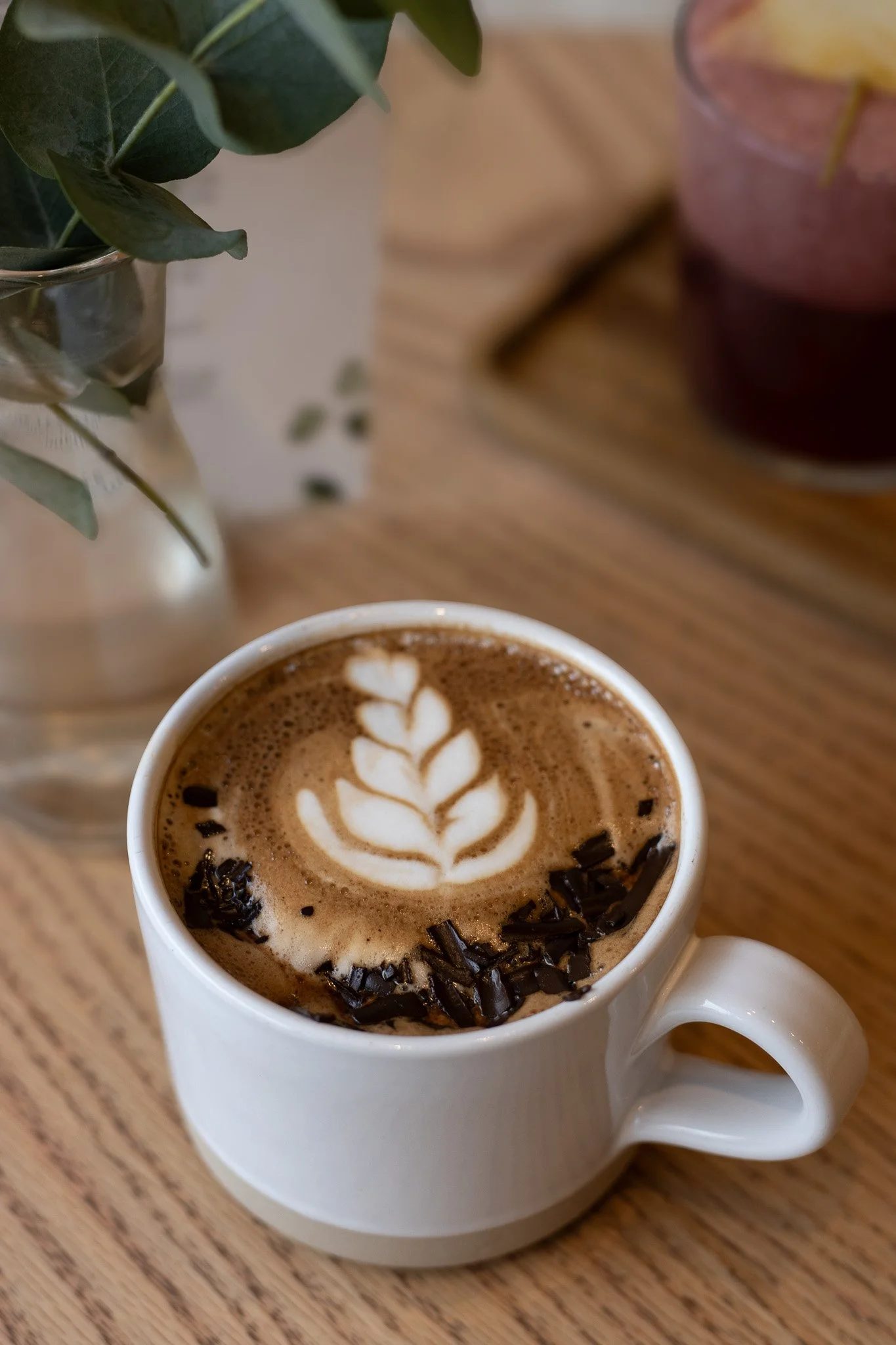 A white mug of hot chocolate with latte art and chocolate shavings, placed on a wooden table, with a vase of green leaves on the left and a pink drink on the right. The Cotswold Farm.