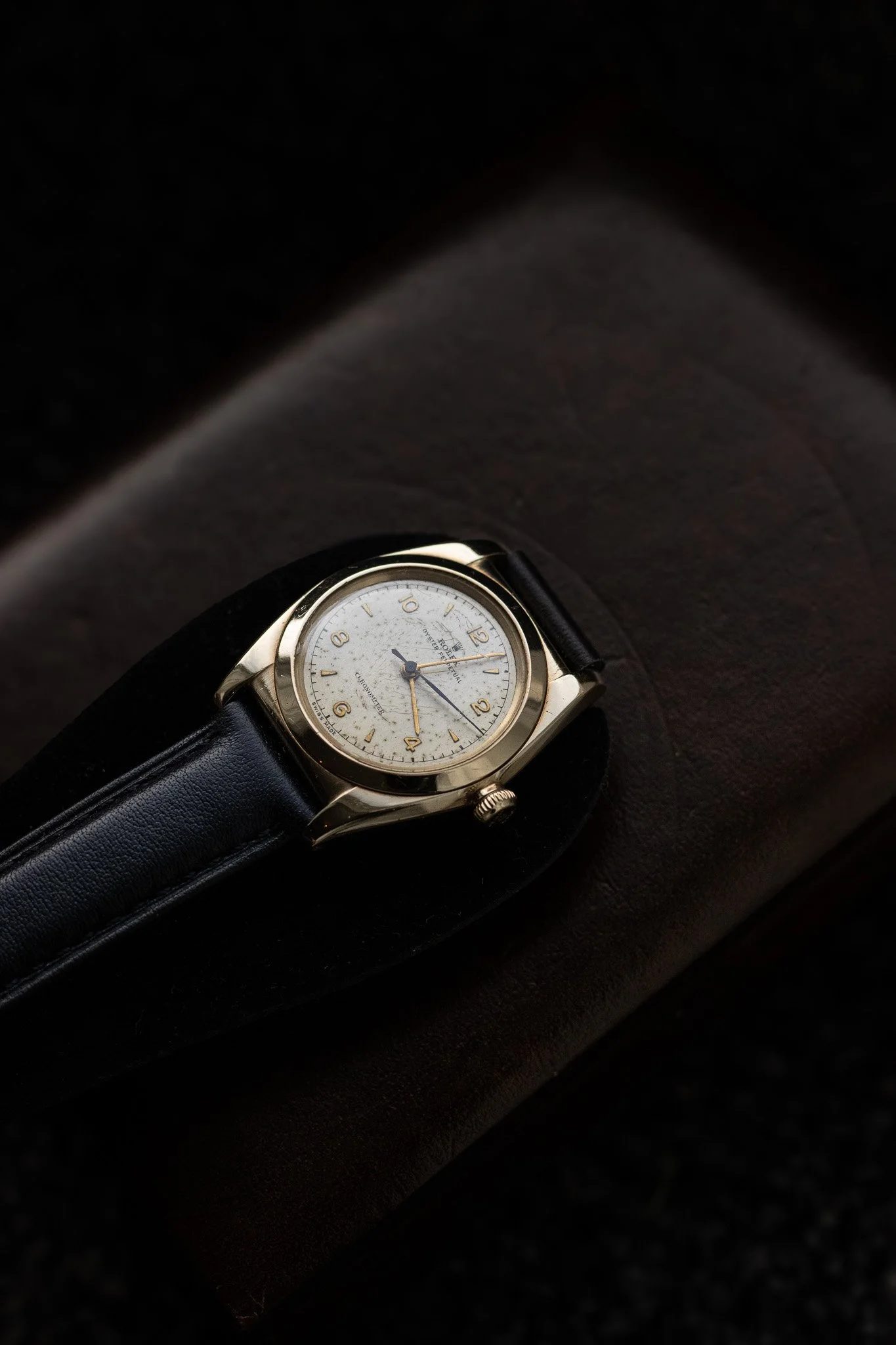 A wristwatch with a light-colored face, gold case, and black leather strap resting on a dark surface. Aurmont. Watches.