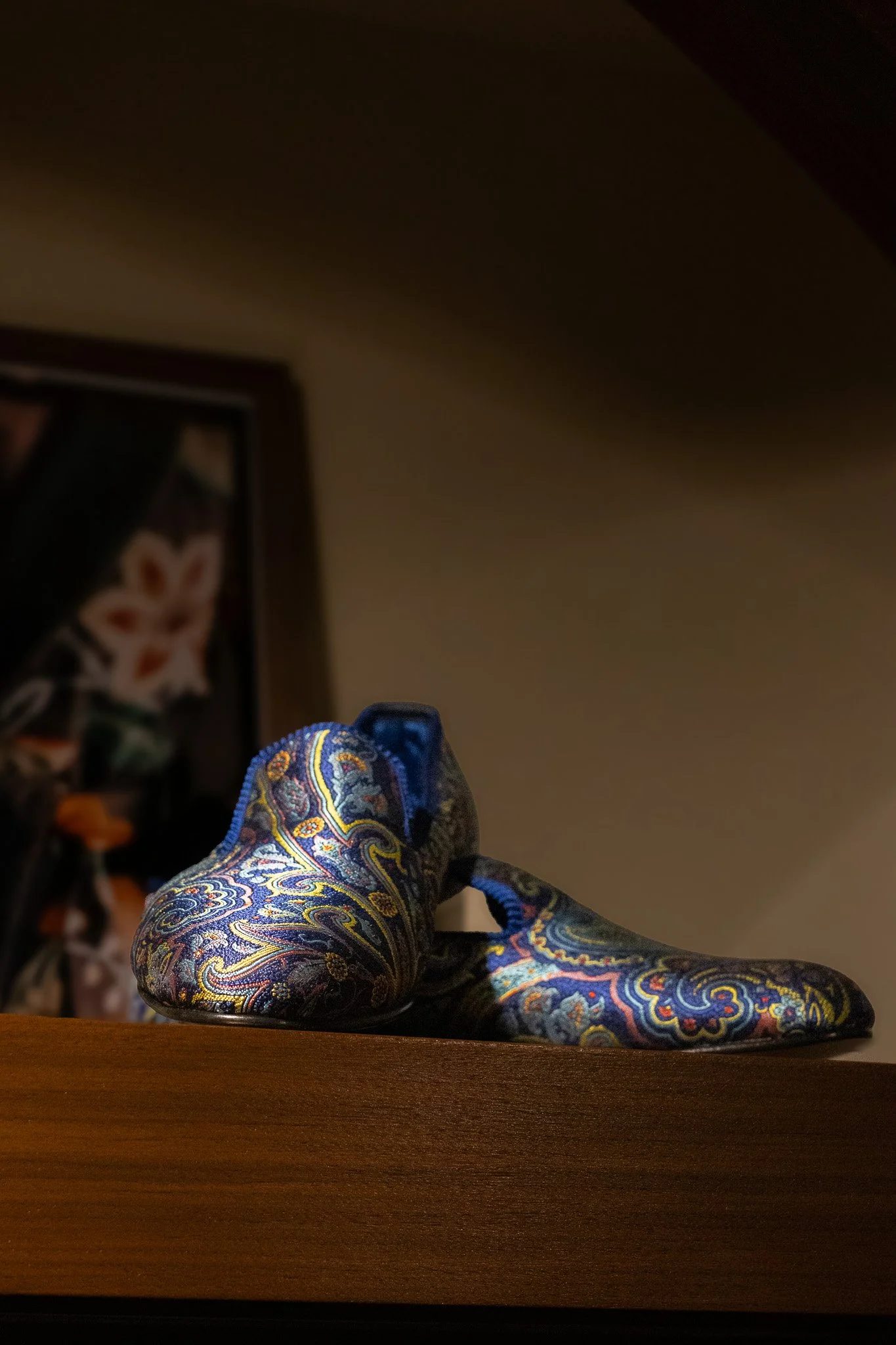 Close-up of a colorful paisley-patterned glass slipper on a wooden surface, with a blurred background. New & Lingwood.
