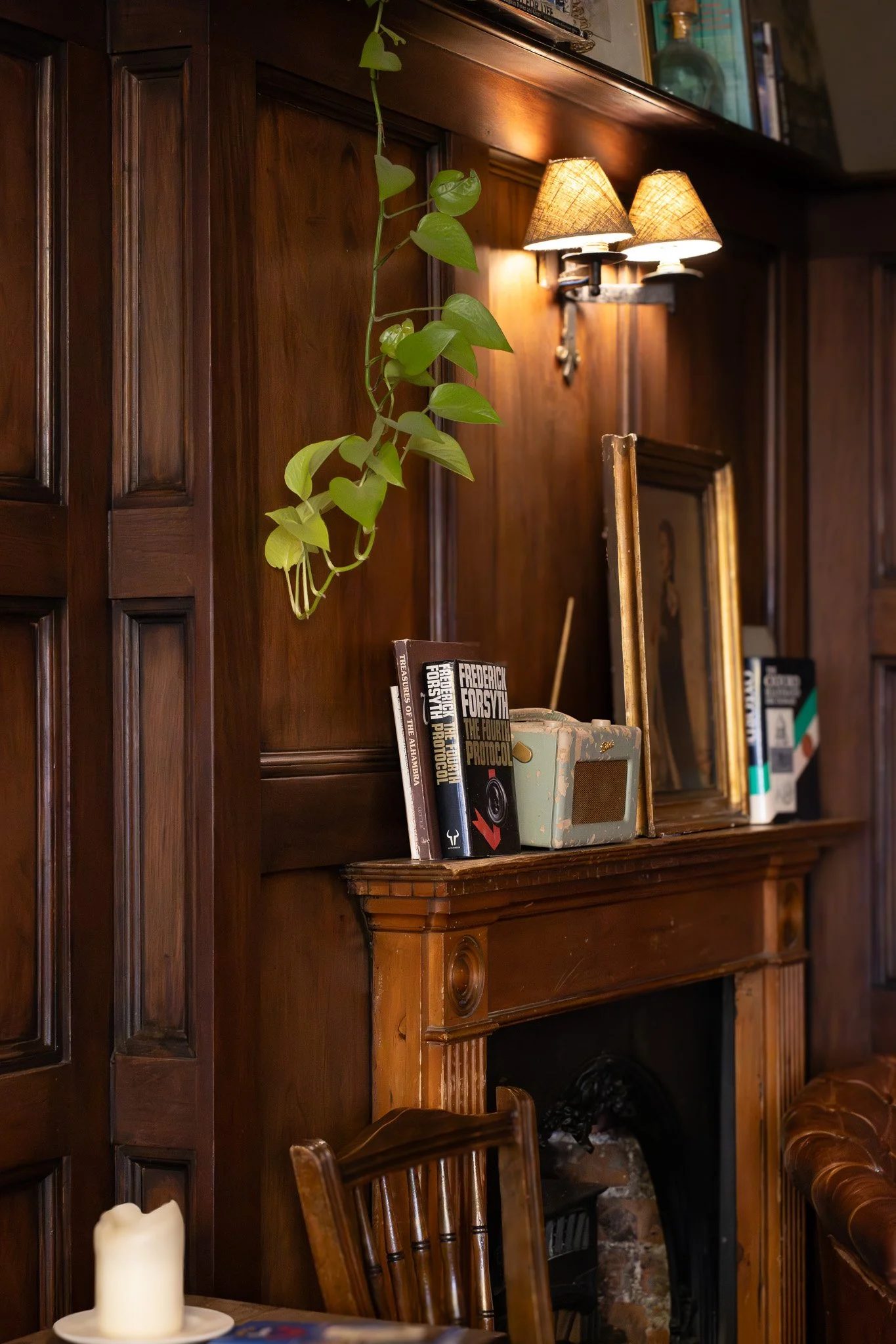 A wooden fireplace mantle and wall, with a green leafy hanging plant, a small bookshelf with books, a mirror, and a wall sconce with two lamps. Part of a table with a candle and a chair are visible in the foreground. The Wilton Arms.