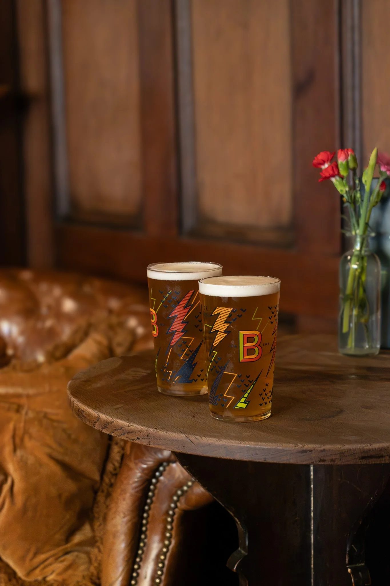 Two glasses of beer on a wooden table with a leather chair and a vase of red flowers in the background. The Wilton Arms.
