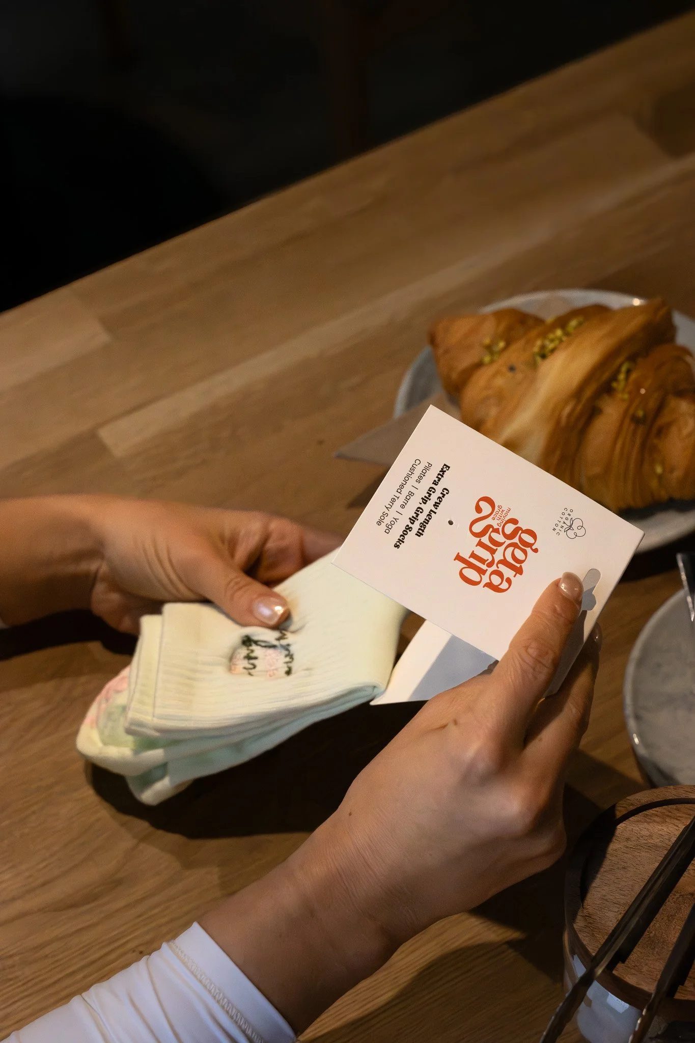 Hands holding a small financial booklet or receipt while sitting at a wooden table, with a croissant and a piece of paper nearby. GetAGrip Apparel.