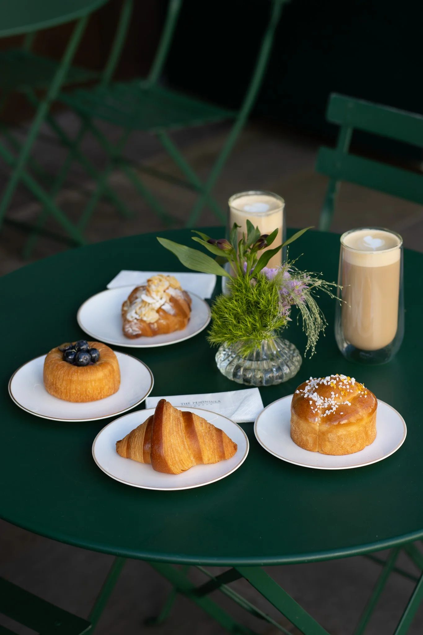 A green round table with assorted pastries, two tall glasses of creamy beverages, and a small floral arrangement in a glass vase. The Peninsula.